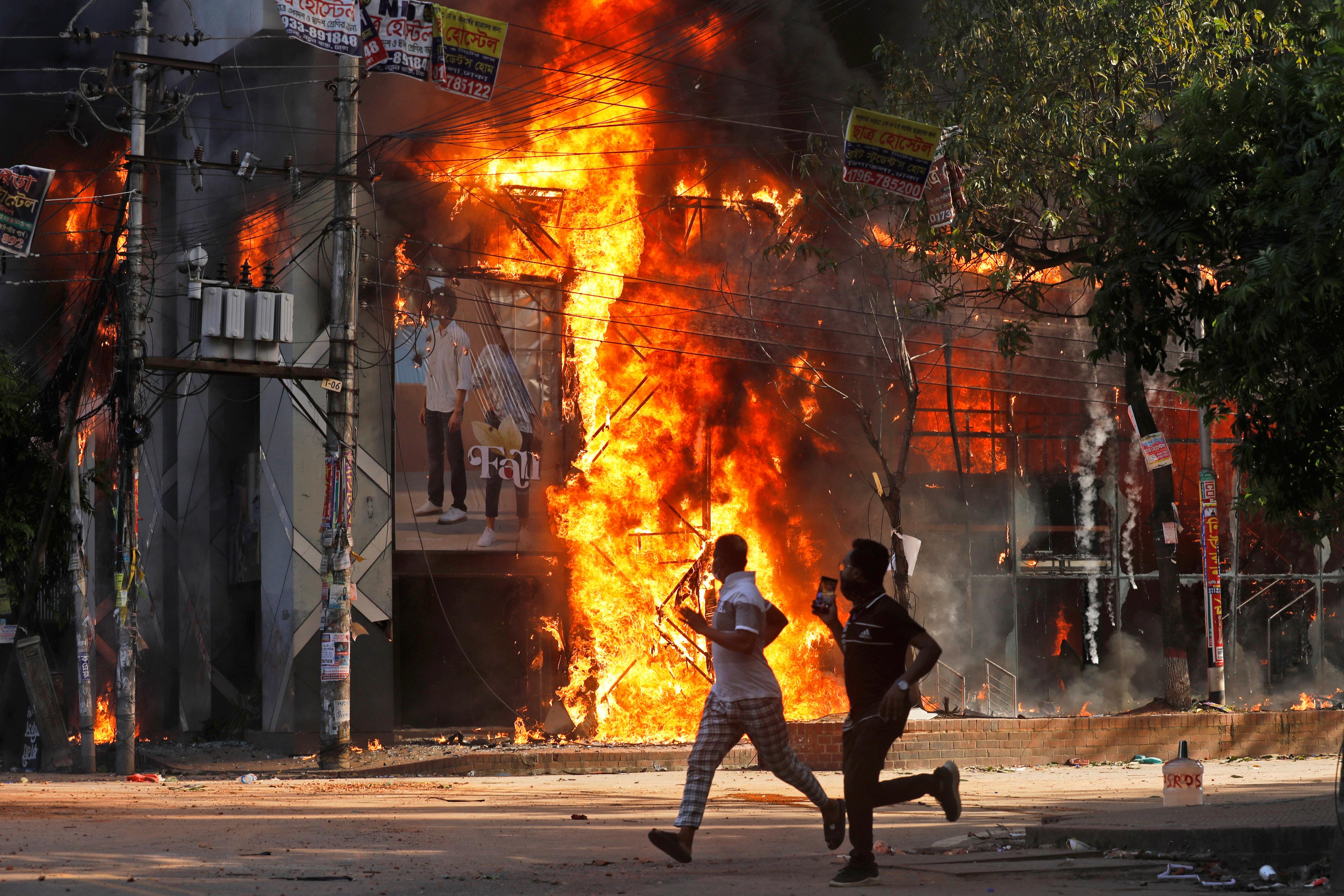 Män springer förbi ett köpcentrum som sattes i brand under en demonstration i huvudstaden Dhaka under söndagen. Foto: Rajib Dhar/AP/TT